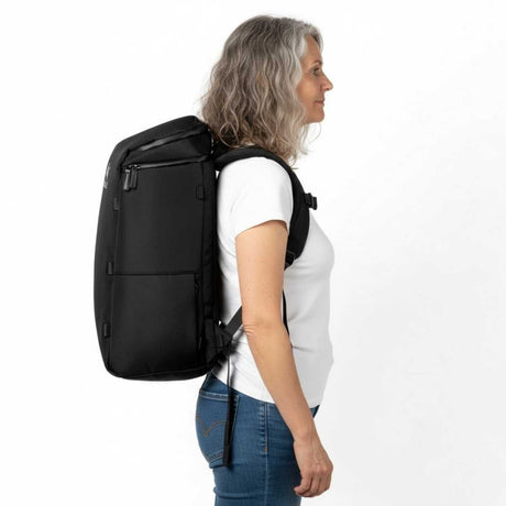 Eldre kvinne i hvit t-skjorte og jeans står i profil med en svart ryggsekk på ryggen.