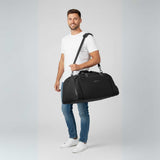 Mann i hvit t-skjorte og jeans bærer en svart duffelbag på skulderen.