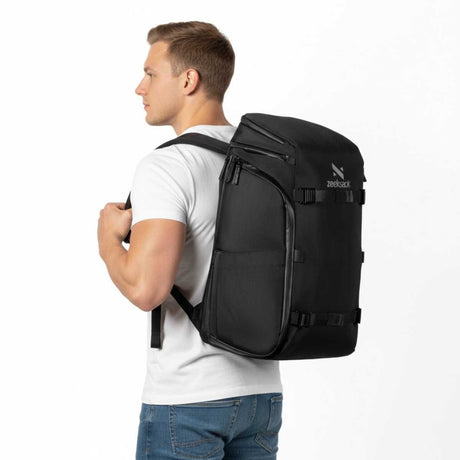 Mann i hvit t-skjorte og jeans står i profil mot høyre med en svart ryggsekk på ryggen.