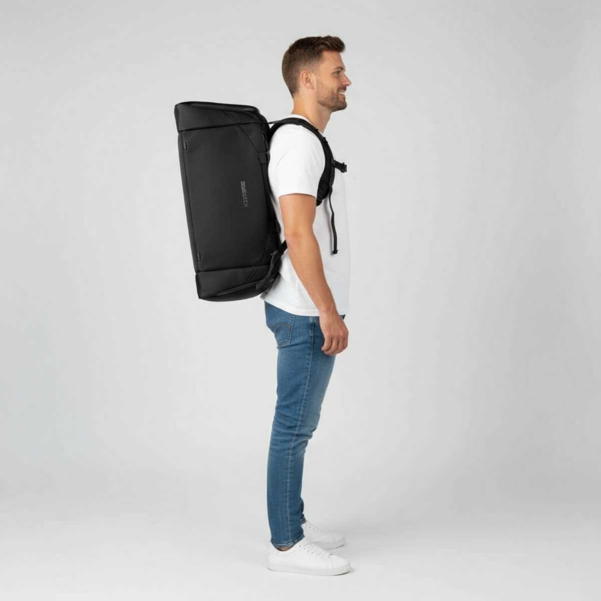 Mann i hvit t-skjorte og jeans står i profil med en svart ryggsekk på ryggen.