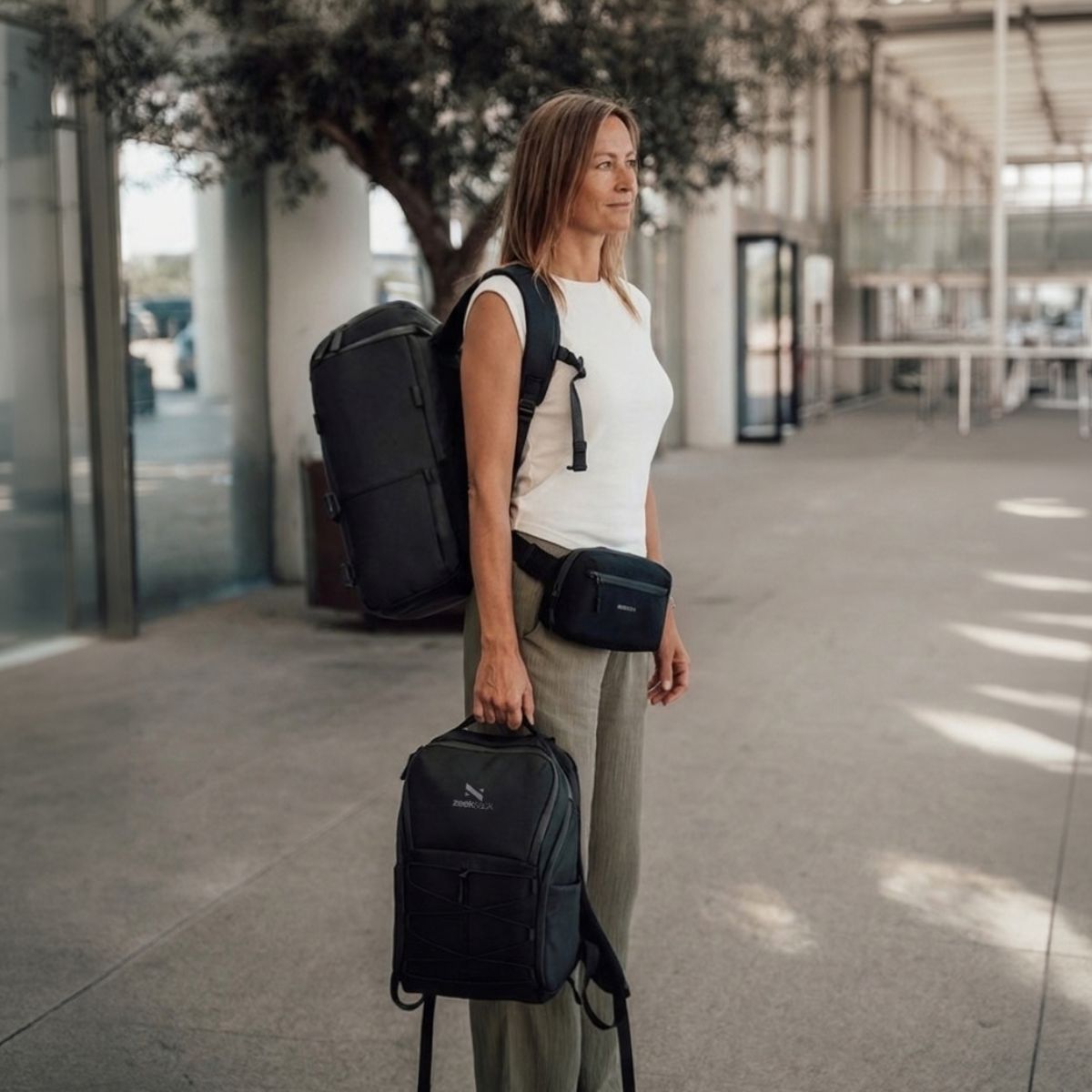 Zeeksack - Kvinne med The Organizer 30L på ryggen, The Underseat 15L i hånden og Crossbody Organizer rundt livet.