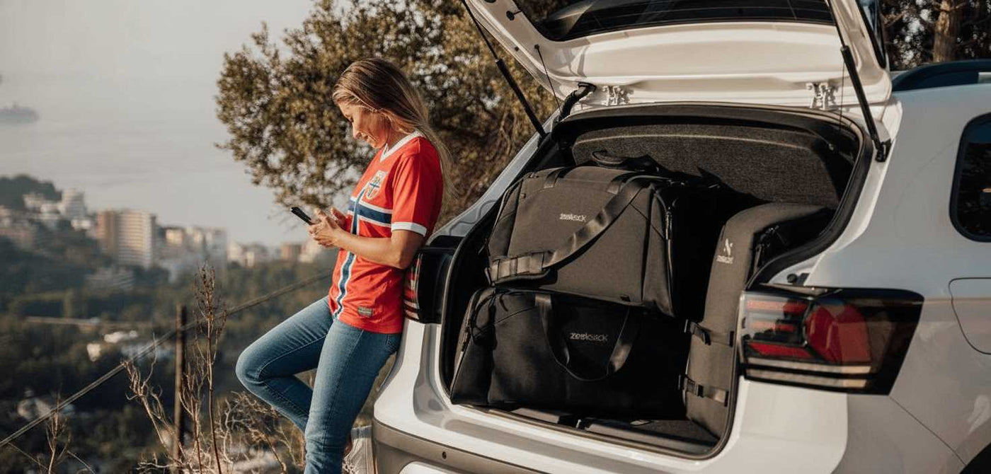 En kvinne i blå jeans, hvite joggesko og en rød trøye i norsk landslagsstil lener seg mot en bil med åpen bagasjeluke. I bagasjerommet ligger svarte sportsbager og en sekk fra Zeeksack. I bakgrunnen ser man utsikt over by og hav.