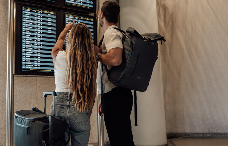 Par med Zeeksack-ryggsekk og koffert sjekker avgangstavle på flyplassen.